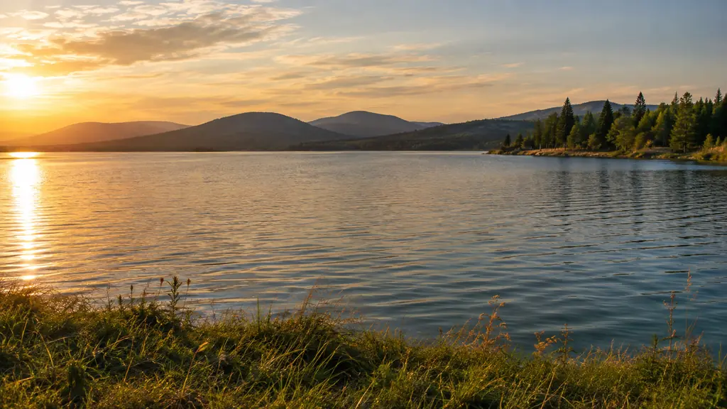 A calm lake at late afternoon seen from a bank, with hills and pines in the distance